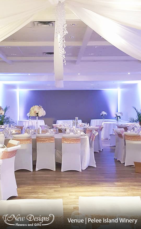 A hall is decorated with a gorgeous draped white ceiling, tall centerpieces of Hydrangeas and roses compact atop a tall clear fluted vase run through with twinkle lights. And ambient lighting spikes upwards in the corners of the room.